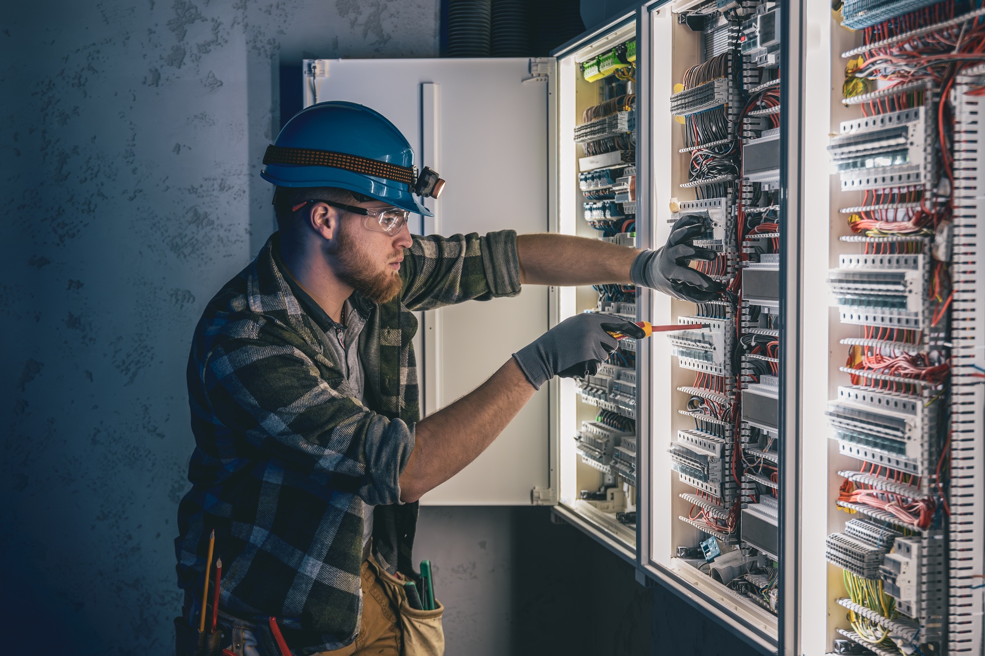 A male electrician works in a switchboard in overalls against the backdrop of emergency lighting.
