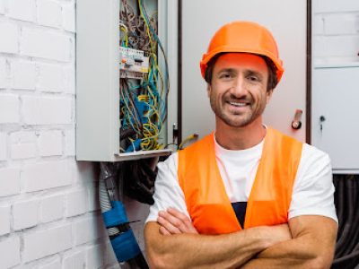 Handsome electrician with crossed arms smiling at camera near electric panel